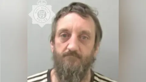 A police mugshot of a man with brown hair and a long brown beard with grey streaks. He is wearing a black and white striped top.