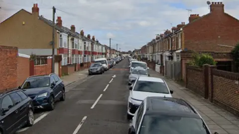 Hollam Road is a narrow one-way street with terraced two-storey houses and cars on both sides of the road.