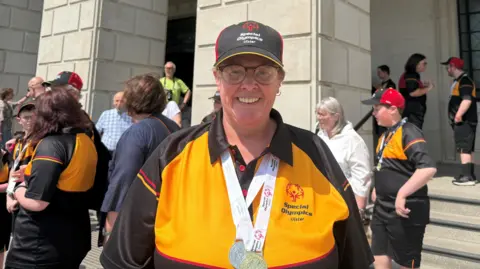 Jackie Stewart. She is wearing a baseball cap, wearing a yellow and black jersey and two medals around her neck.
