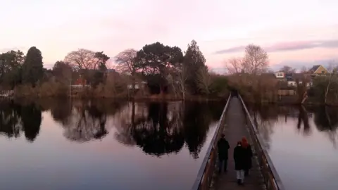 Elsie from the block A wooden footbridge with three people crossing. Part of the sky is purple, which is reflected in the water.