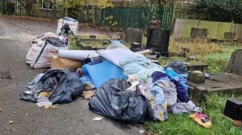 A pile of rubbish is strewn over several graves that have dark or black-coloured headstones. The piles include black bin bags, clothing, cardboard boxes and furniture.