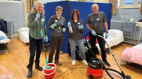 King's Arm Project Four volunteers standing in a room looking at the camera, holding cleaning items by a vacuum cleaner and mop. There are beds around them, and a blue screen. The room has blue walls, windows, with curtains. 