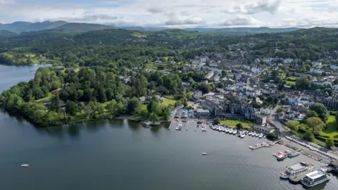 An aerial view of Bowness-on-Windermere, a lake shore town in the Lake District National Park. There are lots of boats moored in the marina and the town stretches out beyond it, surrounded by trees. In the distance are hills and mountains."