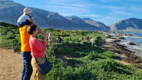 Family photo A side-on photo of Mr Teoh's family standing at the shoreline, facing the sea, with dramatic mountains in the background. There is green shrubbery on the beaches.
