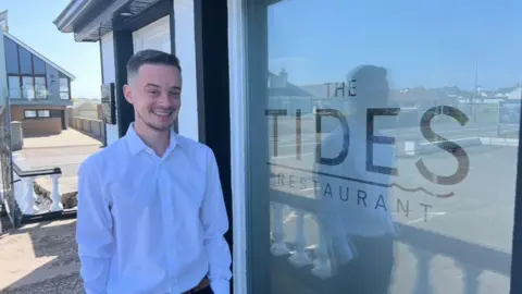 Man wearing a white shirt smiling at the camera. He has dark hair and is standing next to a sign that says Tides