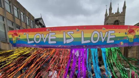A rainbow-coloured banner has the words "love is love" printed on it and there are streamers coming from the bottom of the banner. A tower can be seen in the background.