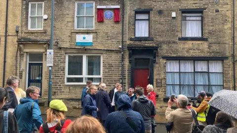 A crowd of people outside a terrace row of offices with a Blue Plaque on the outside wall on the first floor.