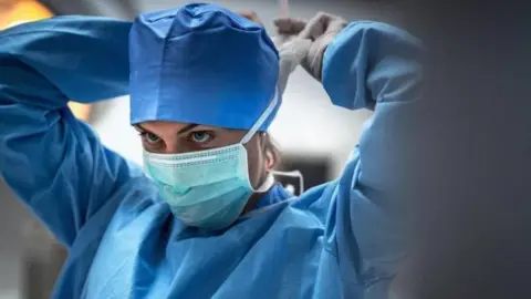 Getty Images A surgeon in blue scrubs putting on a white face mask