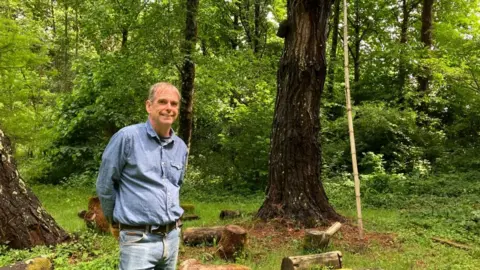 Martin Crawford, dressed in blue jeans and a blue denim shirt, standing by a fully grown tree and some cut logs, in the Forest Garden he created on the Dartington Estate