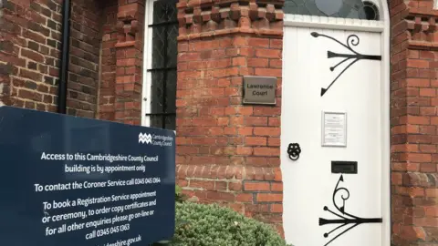 The entrance to Cambridgeshire Coroners' Court, which is a brick building with a white door. There is a black sign outside that offers information to visitors.