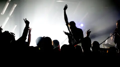 Getty Images Clubbers holding their arms in the air are pictured in silhouette as they watch a band perform, with a guitarist also in silhouette holding his right arm in the air. The stage is backlit.