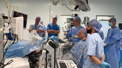 A group of surgeons are standing in a hospital room looking a a screen. They are all in blue protective clothing and have their heads covered.