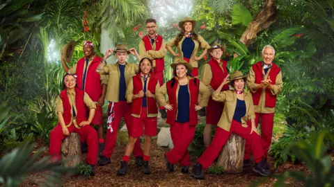 ITV 10 people can be seen wearing various outfits of red and khaki green, with some wearing large khaki hats. They are posed in front of a jungle backdrop as part of the announcement for the new cast of I'm a Celebrity...Get Me Out of Here!