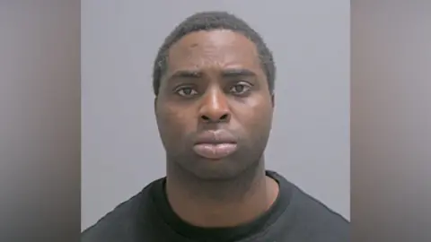 A police custody photo of a man with short dark hair and a black T-shirt.