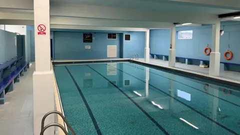 John Devine/BBC A swimming pool, with no-one in or around the water. The building has sky blue walls and darker blue bench seating.