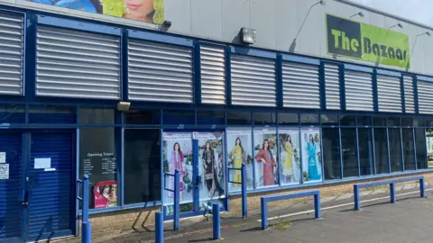 Grace Wood/BBC The front of a market building with glass windows and blue frames. The doors are closed. There are posters in the window showing women in colourful outfits