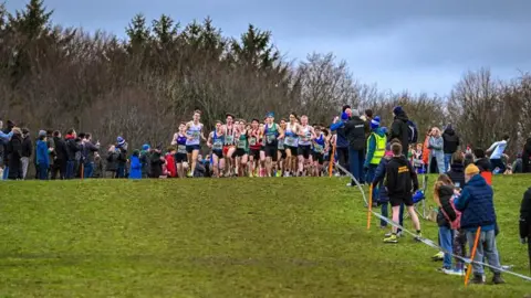 Ben Cuthbertson Male runners coming over the brow of the hill in a tight group. Spectators line the side behind some tape.