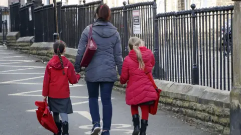 PA A woman in a grey coat and blue jeans with a red hand bag, walking along the road holding the hand of two young girls in red coats and red bags. The woman has dark hair along with the girl on the left, whilst the girl on the right has blond hair. They have their backs to us and are walking adjacent to a row of black school gates which are in a small wall.
