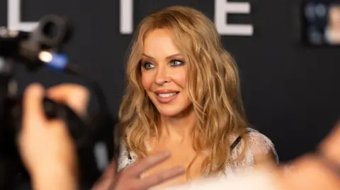 A woman (Kylie Minogue) with wavy blonde hair smiles while being photographed at an event, with blurred cameras and people in the foreground.