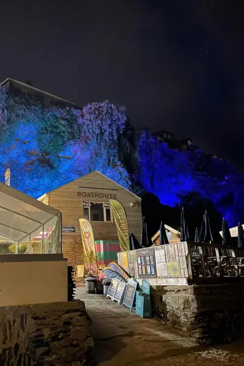 Newquay BID The ivy-covered wall of Newquay Harbour is lit up with blue and purple lights. In the foreground is a building with the letters BOATHOUSE on it, two flags advertising Cornish Rattler and a row of wooden chalk board signs and signs above promoting businesses in the harbour. On the left is a marquee and on the right is a raised area covered in wooden chairs.