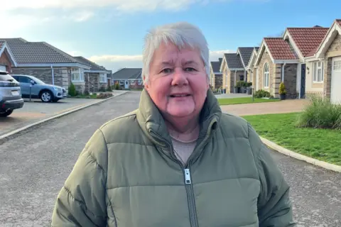 Matt Knight/BBC A lady in a green coat stands in the middle of a road. There are new-build bungalows either side of her.