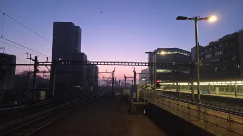 DereksDisco A purple sky over a train station. There are power lines over the tracks. The platforms are illuminated with artificial light