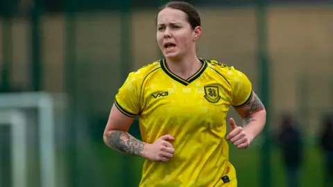 NashyPhoto A head and shoulders of a female footballer wearing a yellow football top while mid-run during a game.