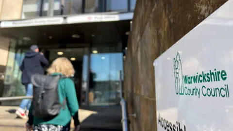 The entrance to Warwickshire County Council's headquarters is photographed from outside. The entrance is out of focus in the background as two people enter, in the lower right foreground a sign displaying the Warwickshire County Council logo with bear and staff is clearly visible.