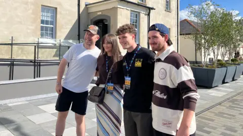 Member of Kingfishr taking pictures with fans. From left to right, a man is wearing a cap, white shirt and black shorts; a girl has long brown hair with a fringe, wearing a black t-shirt, striped skirt, black handbag, and an access all areas lanyard; a man with brown hair, wearing a black t-shirt, black trousers and an access all areas lanyard; a mad wearing a blue backwards cap and brown and white rugby top.