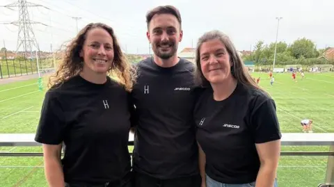 LooseHeadz Amber Reed, left, has long curly brown hair. She stands next to a man with dark brown hair. On his right is a woman with long brown straight hair, The trio are wearing black T-shirts and are standing in front of a rugby pitch.
