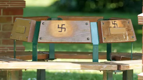 A hexagonal bench made from wood and green metal. There are swastikas carved into the back rest of two of the benches.