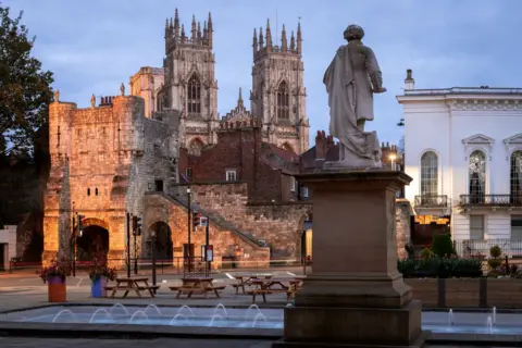 Getty Images Jets of water fire into a pool at the base of a statue of a man stood on a plinth. In the background the towers of York Minster can be seen.