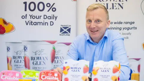 A man in a blue shirt with short blond hair sits behind yoghurt products with the Rowan Glen name on them