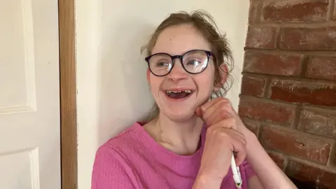 A young woman wearing round black glasses and a pink top is photographed smiling as she looks off to the left of the camera. She is sat down with a brick wall behind her.