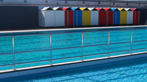 Hilsea Lido A light blue pool with changing rooms in different colours in the background.
