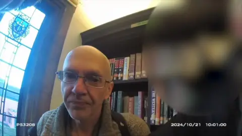 Roy Barclay captured on bodycam footage in Ipswich library, sat in front of  a book case with a window nearby