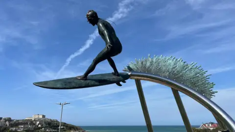 A statue of a surfer on a surf board suspended on a steel pole with the sea behind it. Behind the surfer is a bronze wave symbol.