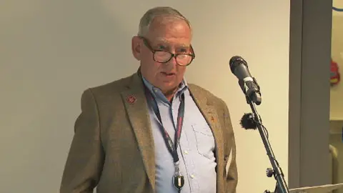 BBC Tony Dawson stood in front of a microphone and lectern. He is wearing a blue shirt, lanyward and suit jacket. He is wearing glasses and looking forward.