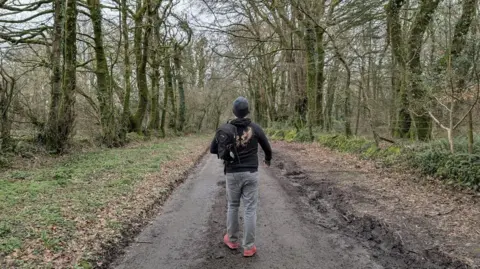 Dean Cooper Dean Cooper on a walk in Cornwall which helps him deal with burnout and stress. He is seen walking into a forest with a backpack.