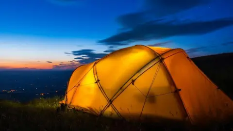 A yellow tent lit from inside. It is on a patch of grass. Lights can be seen in the distance. The sky is blue and orange with some clouds.