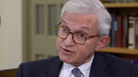 Sir Geoffrey Vos has short gray hair and wears glasses. He is wearing a pinstripe navy suit with a white shirt and tie. There is a bookshelf behind him