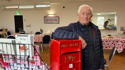 BBC Nick Gibbs - dengan jaket biru dan rambut abu -abu - berdiri di dalam aula komunitas yang meninggalkan kotak pos merah tradisional. Stand of Valentine Day Cards ada di sebelahnya, dan di belakang adalah kafe, yang termasuk meja kue dengan taplak meja bertema hati.