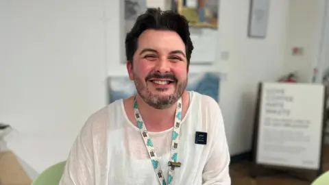 Jack Maclean/BBC Jack Baxter is sitting down at a desk on a green chair. He is looking directly at the camera and smiling. He has a white, floaty long-sleeved T-shirt on and a lanyard around his neck.