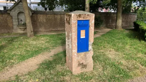 A blue post box set inside a damaged concrete surround with grass, trees, and a stone wall behind it.