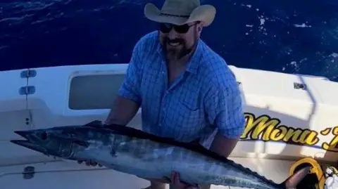 Patrick Collins Patrick Collins. He has a dark beard, wearing a stetson hat, sunglasses and blue check shirt. He is on a speedboat holding a large fish.