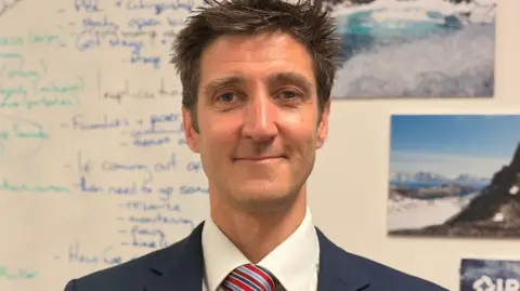 Phil Avery, director of education at Bohunt school in front of a white board. He has short brown hair and is looking at the camera smiling. He has a blue jacket with a white shirt and a red and blue stripped tie. Behind him the board has out of focus blue writing. There is a photo of what appears to be a glacier and a mountain.