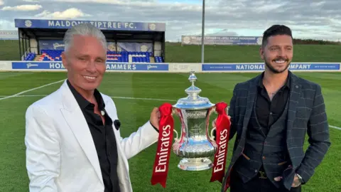 Warren Muggleton/BBC Barrie and Scott Drewitt-Barlow are holding the FA Cup - which is a silver trophy with red ribbons - on the pitch at Maldon and Tiptree FC. Behind them is a small blue stand. Barrie is wearing a black shirt and a white blazer. Scott is wearing a checkered dark grey suit.