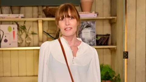 A woman wearing a cream blouse with shelves in the background filled with pots, plants and books