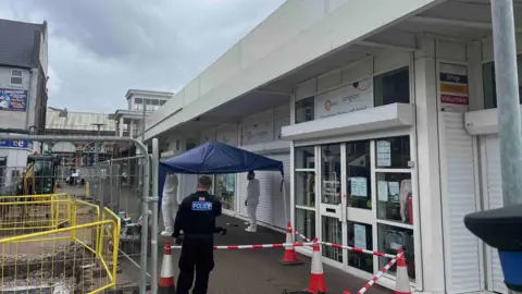 A police officer wearing black with a blue "police" sign on his back. There are other people, who look like forensic officers, in shot and a red and white cordon with cones.
