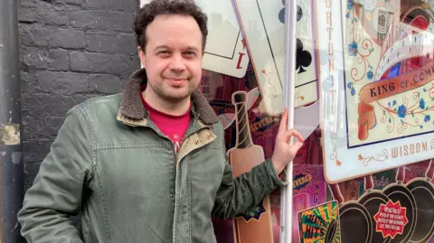 A man wearing a green jacket and red top stands smiling next to a brick wall, and leans on the old shop window with part of the mural visible behind him. The design includes a cricket bat, and playing card and writing. 